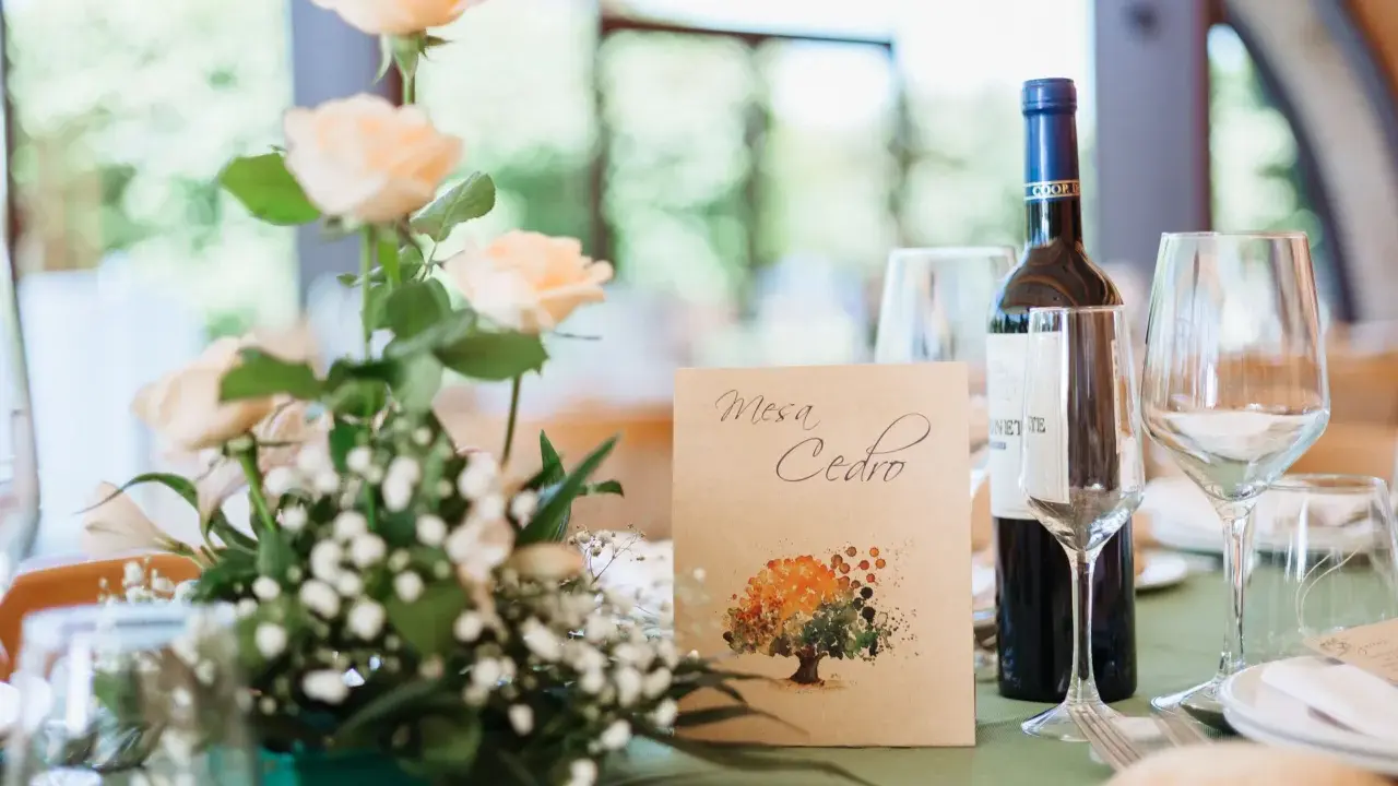 Detalle de mesa de boda con flores color melocotón, botella de vino y tarjeta con árbol pintado. Ideas originales para bodas.